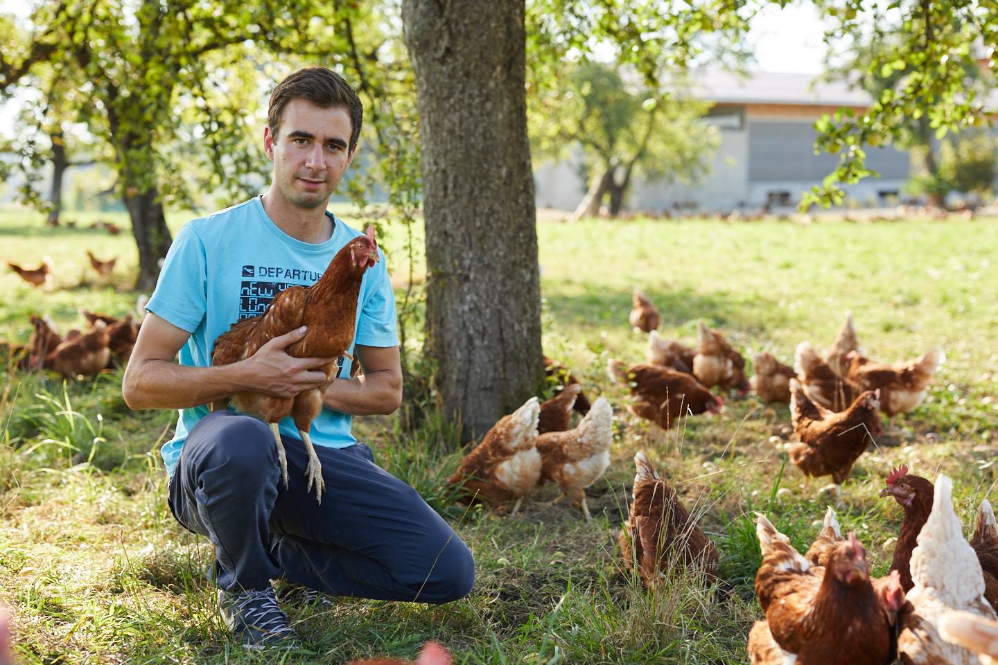 Gebhard Kröss Sitzt Auf Der Wiese Mit Einem Huhn In Der Hand, Im Hintergrund Sind Außerdem Noch Andere Hühner Sichtbar