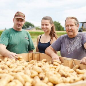 Familie Fink Steht Hinter Einem Anhänger, Der Mit Kartoffeln Gefüllt Ist Und Lächelt Dabei In Die Kamera