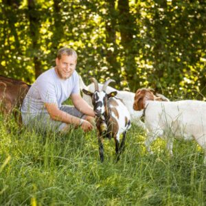 Norbert Schedler Sitzt Neben Seinen Ziegen In Der Wiese Und Lächelt In Die Kamera, Im Hintergrund Sind Mehrere Bäume Erkennbar