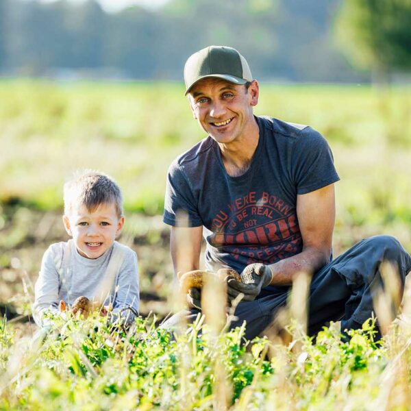 Thomas Bischof kniet neben seinen Sohn in einem Kartoffelfeld, beide halten Kartoffeln in den Händen und lächelln in die Kamera.