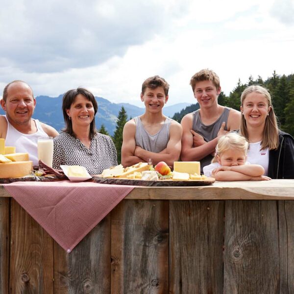 Die Familie Meusburger steht hinter einer Holzbar auf dieser herhafte Käseplatten der Alpe Gemeine Brongen aufgetischt sind.