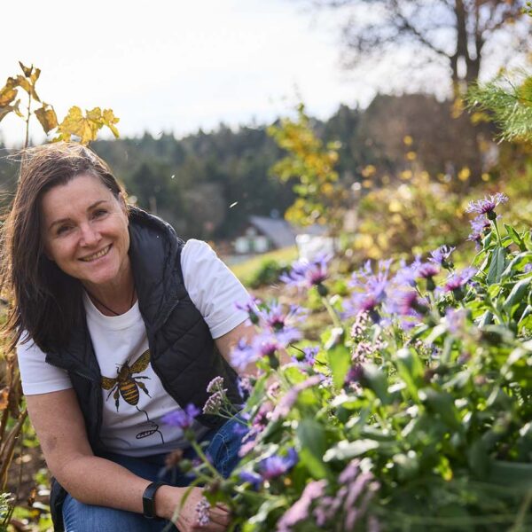 Martina Pinzger kniet zwischen Blumen in ihren Garten und lächelt in die Kamera.