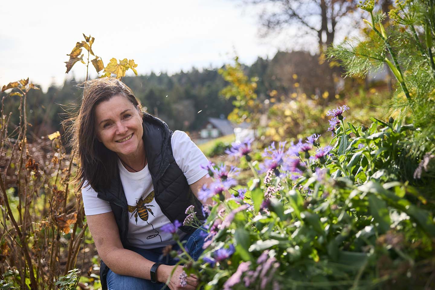 Martina Pinzger Kniet Zwischen Blumen In Ihren Garten Und Lächelt In Die Kamera.