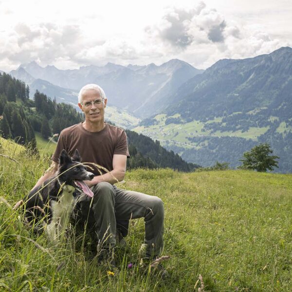 Andreas Burtscher sitzt mit seinem Hund in einer Bergwiese in St. Gerold und lächelt in die Kamera.