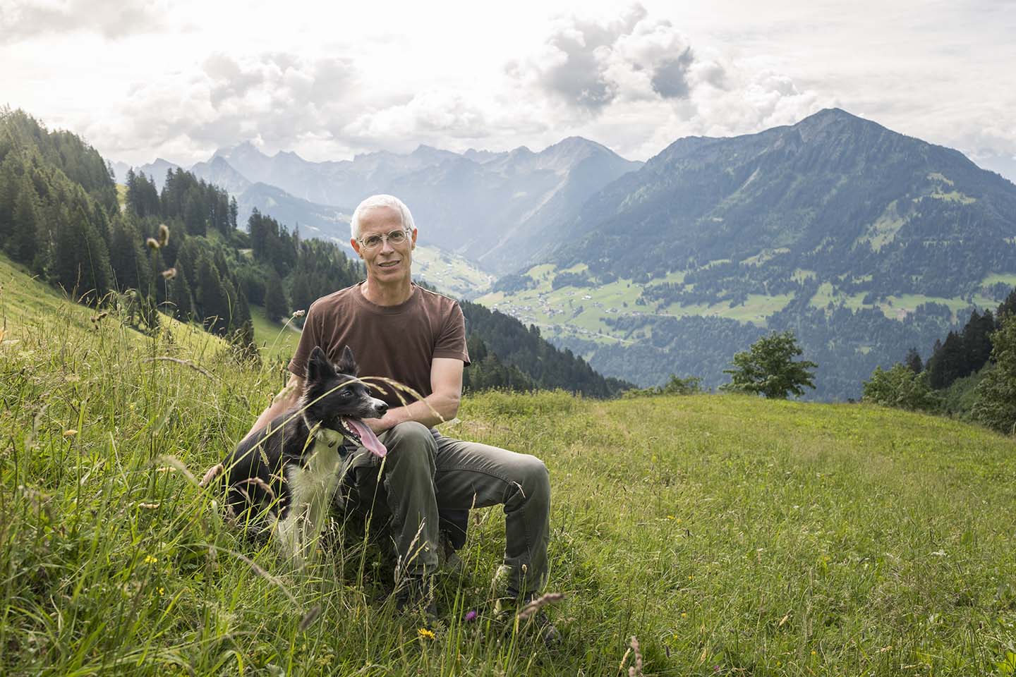Andreas Burtscher Sitzt Mit Seinem Hund In Einer Bergwiese In St. Gerold Und Lächelt In Die Kamera.